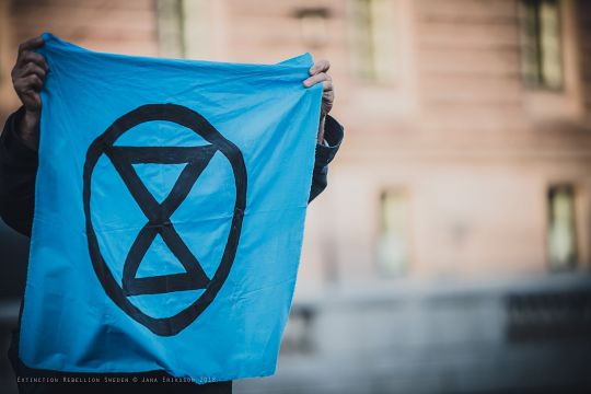 Ein himmelblaues quadratisches Transparent mit einem Extinction-Rebellion-Logo, einer stilisierten Sanduhr, wird ins Bild gehalten.