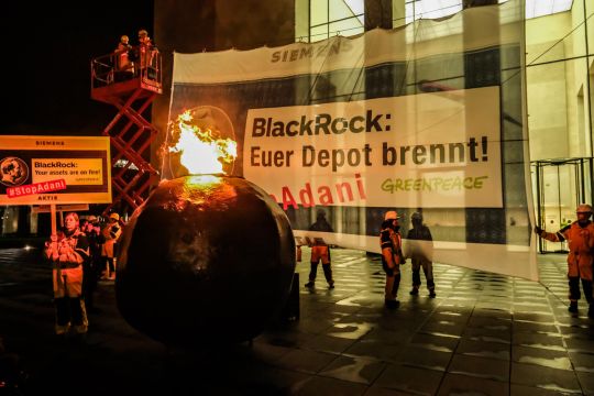"Blackrock: Euer Depot brennt!", steht auf einem großen Siemens-Aktien-Banner am Gebäude der Blackrock-Zentrale, davor brennt ein großer schwarzer Stein.