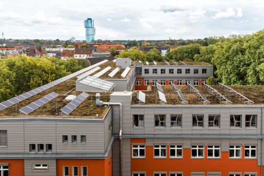 Auf dem niedrig begrünten Flachdach eines mehrstöckigen Gebäudes sind Solarpaneele schräg aufgestellt, am Horizont ist das Bochumer Exzenterhaus zu sehen.