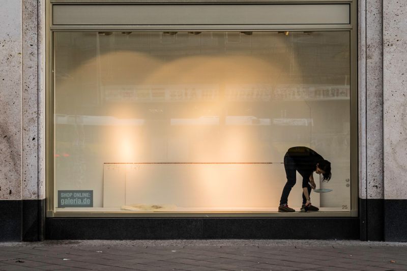 Leeres Schaufenster, in dem eine Angestellte aufräumt, auf einem kleinen Schild steht: Shop online: galeria.de.