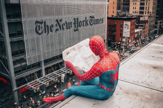Mensch in Spiderman-Kostüm liest auf dem Dach Zeitung, gegenüber sieht man das Gebäude der New York Times