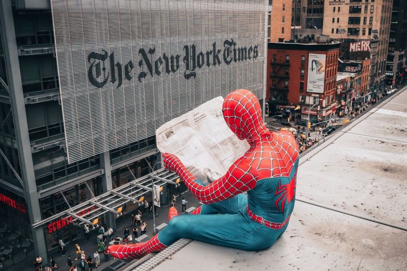 Mensch im Spiderman-Kostüm liest auf dem Dach Zeitung, gegenüber sieht man das Gebäude der New York Times.