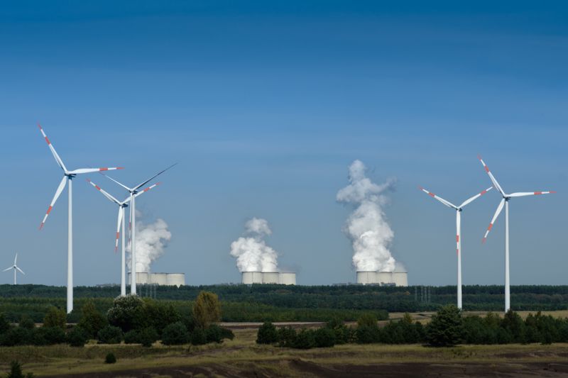 Einige Windräder in Landschaft mit Wäldern und feldern, im Hintergrund ein großes Kohlekraftwerk mit neun dampfenden Kühltürmen.