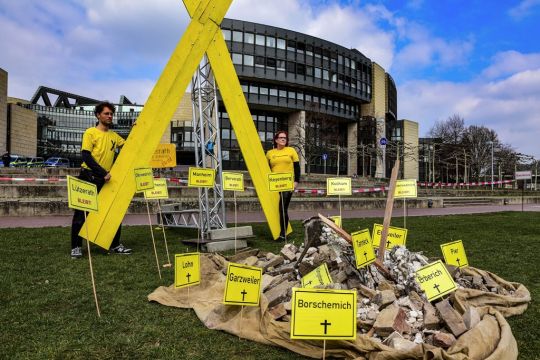 Zwei demonstrierende Personen mit großem gelbem X, Ortsschildern abgebaggerter oder bedrohter Dörfer und einem symbolischen Schutthaufen auf der Wiese vor dem Landtagsgebäude in Düsseldorf.