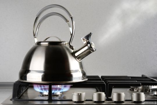 Ein silbrig-metallisch glänzender neuer Teekessel wird auf dem Herd von einer Gasflamme erhitzt, Wasserdampf entweicht und lässt die Flöte ertönen.