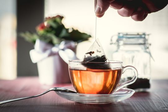 Ein tetraederförmiger Kunststoff-Teebeutel wird aus einer gläsernen Teetasse genommen, der Tee hat die typische, eher noch helle Farbe.