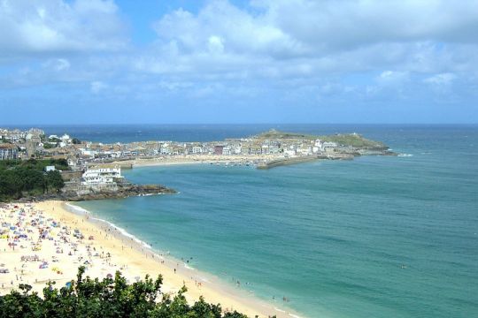 Aufnahme von St Ives, wo der G7-Gipfel stattfindet. Zu sehen sind eine niedrig bebaute Halbinsel, ein breiter Strand, das blaugrüne Meer und ein heiterer Himmel.