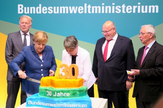 Angela Merkel schneidet eine Torte an, während Barbara Hendricks hilft und Jürgen Trittin, Peter Altmaier und Klaus Töpfer zusehen. Auf der Torte steht: 30 Jahre Bundesumweltministerium.