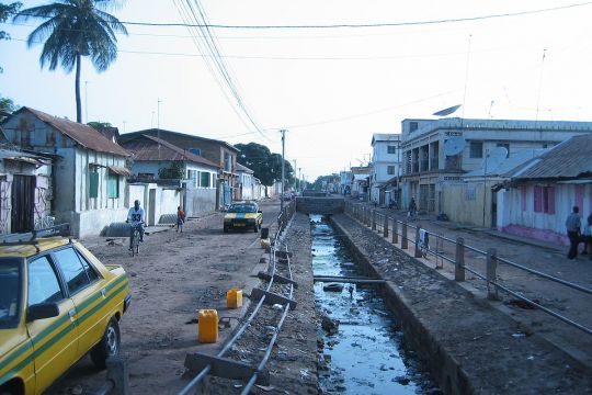 Unbefestigte Straße, in deren Mittelstreifen ein kleiner Kanal verläuft,links und rechts ein- bs zweistöckige Häuser.
