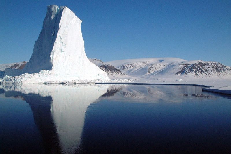 Steil aufragender Eisberg am Ufer vor einer Eiswüstenlandschaft mit flacheren rundlichen Erhebungen.