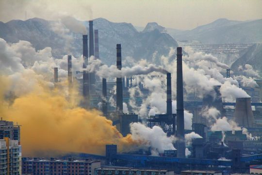 Großer Industriepark im Gebirge, Kühltürme und Schornsteine von Kraftwerken und Industrieanlagen stoßen sehr viel weißen Dampf und orangefarbenen sowie grauen Rauch aus.