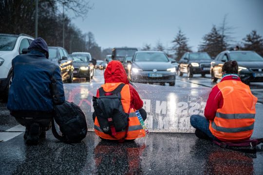 drei Aktivisten blockieren sitzend eine Straße mit zahlreichen Autos