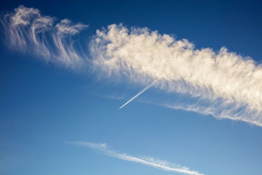 Weißer Zirruswolkenstreifen am blauen Himmel, durch den ein Flugzeug fliegt, von dem nur ein kleiner Kondensstreifen zu erkennen ist.