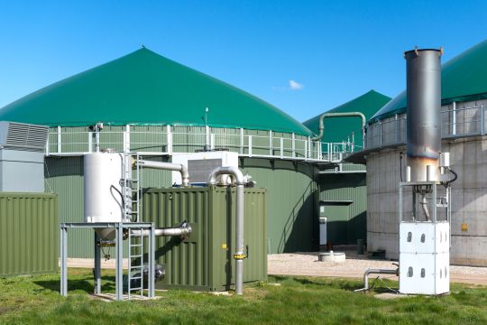 Biogasanlage mit den typischen runden Gasbehältern und einer Notfackel. Das ist ein rund zwei Meter hoher Metall-Schornstein auf einer quaderförmigen Unterkonstruktion, die etwas abseits steht. 