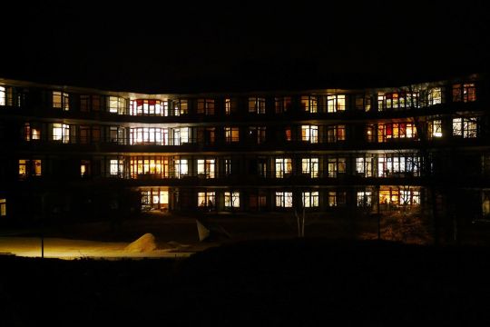 Vierstöckiger moderner Plattenbau im Dunkeln, die meisten Fenster sind beleuchtet.