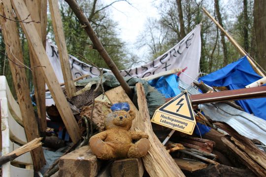 Barrikade aus Holzlatten, -balken und Gerümpel vor Waldbäumen, darauf ein gelbes Warnschild "Hochspannung Lebensgefahr" und ein Teddy-Bär