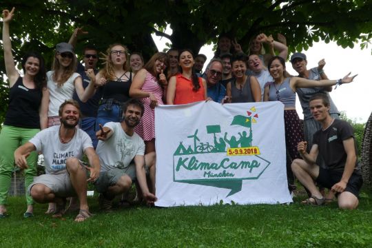 Gruppe lachender und begeistert gestikulierender junger Menschen vor einem Baum, manche halten ein Transparent mit der Aufschrift "Klimacamp München"