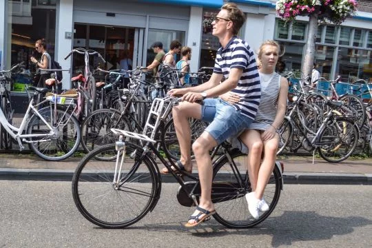 Junges Paar auf einem Fahrrad, sie sitzt quer auf dem Gepäckträger, im Hintergrund viele Fahrräder am Straßenrand.