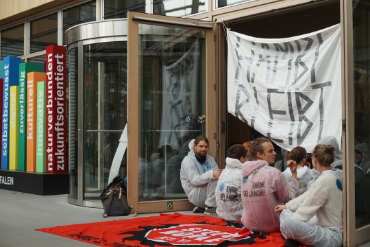 Aktivisten mit dem Banner "Hambi bleibt" sitzen vor einer Tür in der Landesvertretung NRW. 