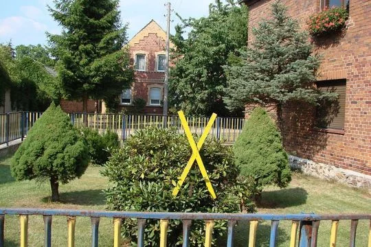 großes, gelbes Warnkreuz aus Holz in einem gepflegten Garten, ringsum dörfliche Backsteinhäuser