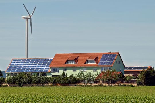 Großes Anwesen auf dem Land mit Windrad und Solardächern, über ein Feld aufgenommen.