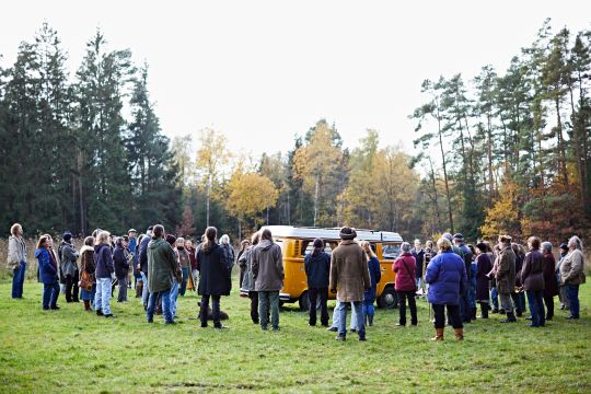 Menschen stehen auf einer Waldlichtung um einen gelben Kleinbus herum.