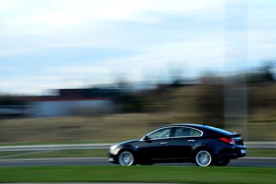 fahrendes schwarzes Auto auf einer Straße, dahinter unscharf Bäume und ein Feld