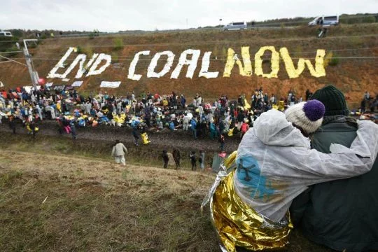 Zwei Menschen umarmen sich im Vordergrund, weiter hinten Menschenmassen bei der Schienenblockade, Schriftzug "End Coal Now" auf Hügel dahinter