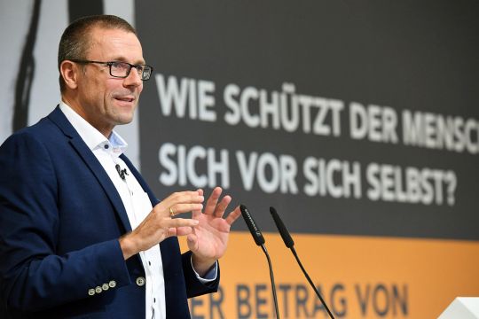 Uwe Schneidewind redet vor einem Bühnenhintergrund mit der Aufschrift: "Wie schützt der Mensch sich vor sich selbst?".