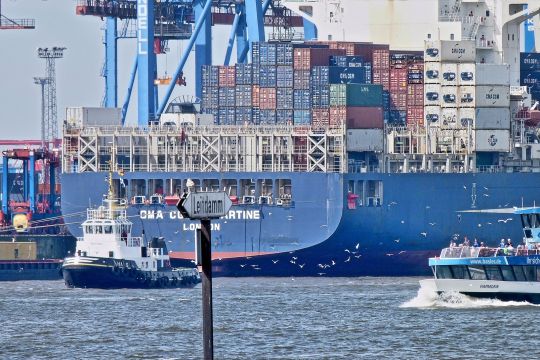 Heck eines riesigen Containerschiffs am Kai des Hamburger Hafens, davor ein Schlepper und ein Ausflugsschiff.