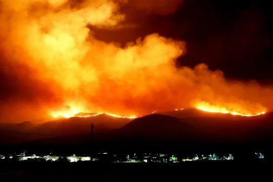 Riesige Flammen in der Nacht über einem Höhenzug im Hintergrund, vorn die Lichter einer Siedlung.