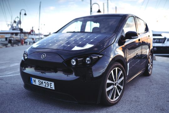 Das Bild zeigt ein schwarzes Elektroauto mit Solarzellen auf der Motorhaube auf einer Straße. 