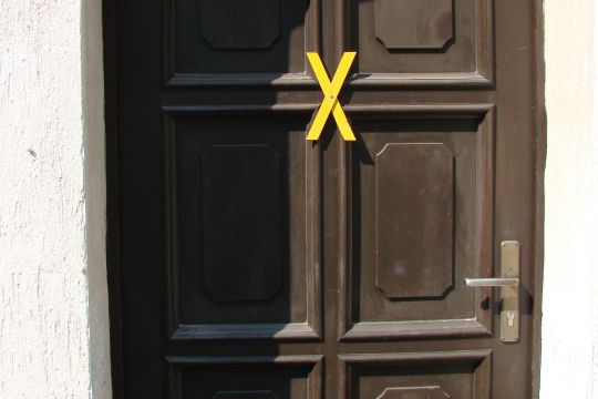 Ein kleines gelbes Kreuz ähnlich einem X ist an eine Haustür aus Holz geschraubt.