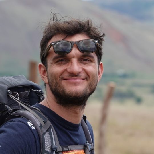 Tobias Baedeker mit hochgeschobener Sonnenbrille und Reiserucksack in bergiger Landschaft