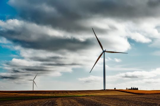 Zwei Windräder stehen auf freiem Feld, am Himmel dunkle Wolken, knapp hinter dem Horizont ein Gehöft.