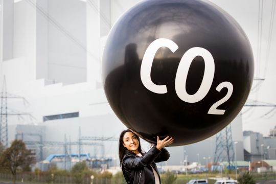 Eine Frau hält einen sehr großen schwarzen Ballon mit der Aufschrift "CO₂" hoch, weiter im Hintergrund ein Kohlekraftwerk.