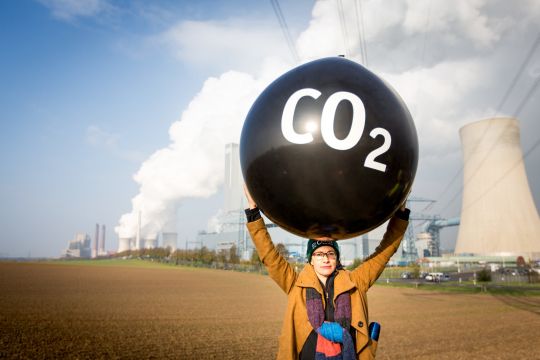 Eine Frau hält einen sehr großen schwarzen Ballon mit der Aufschrift "CO₂" hoch, weiter im Hintergrund ein Kohlekraftwerk.