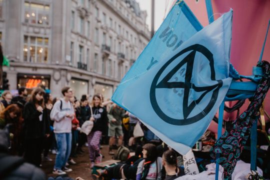 Blaue Flagge mit dem Logo von "Extinction Rebellion" (umkreiste Sanduhr), dahinter unscharf Menschenmasse auf einer großen Kreuzung