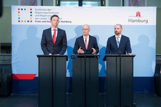 Drei Männer stehen nebeneinander an Rednerpulten vor einem aufrollbaren Hintergrund mit der Aufschrift: "Konferenz der Regierungschefinnen und Regierungschefs der Länder"