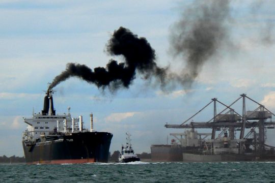 Hochseeschiff mit einer schmutzigen ​Rauchfahne, im Hintergrund Hafenanlagen.
