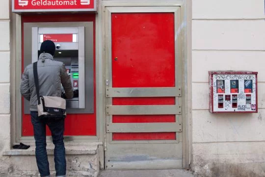 Mann hebt Geld an einem Sparkassen-Automaten ab, der auf der Straße in einem kleinen Ladenschaufenster installiert ist, auch die Tür daneben ist offenbar dauerhaft geschlossen, daneben hängt noch ein Kaugummi-Automat an der grauen Wand.