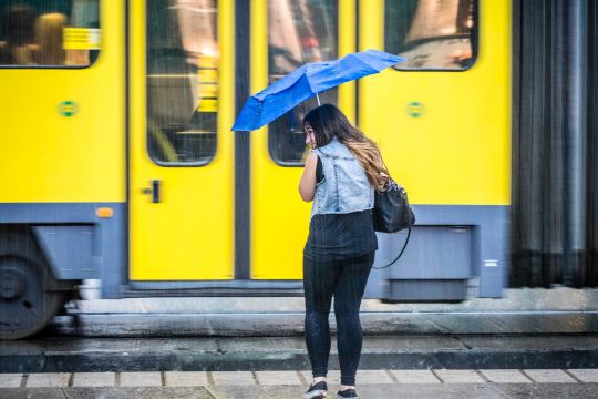 Frau steht im Regen vor einer Straßenbahn, ihr blauer Regenschirm wird vom Wind gebeutelt.