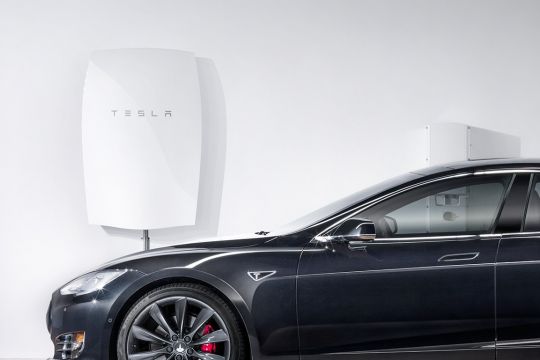 Ein weißer Solarstrom-Hausspeicher "Tesla Powerwall" hängt an der Wand, davor steht ein schwarzes E-Auto vom Typ Tesla Model S. 