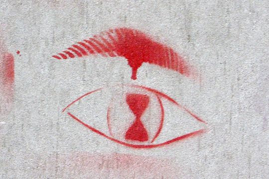 Ein rotes Graffito auf grauer Wand zeigt ein Auge, dessen Pupille wie eine Sanduhr aussieht.