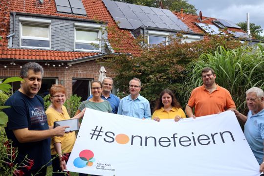 Acht Leute stehen vor einem Mehrfamilienhaus mit Solaranlage und halten ein Transparent mit der Aufschrift "Sonne feiern".