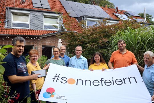 Acht Leute stehen vor einem Mehrfamilienhaus mit Solaranlage und halten ein Transparent mit der Aufschrift