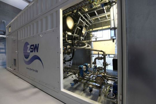 Hinter der geöffneten Tür eines weißen Containers mit der Aufschrift "ZSW" sind ein Kessel und technische Apparaturen zu sehen.