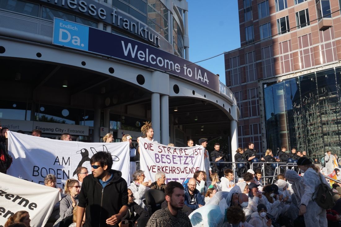 Protesierende sitzen vor einem Messegebäude, im Hintergrund ein Schild 