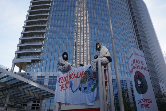 Mehrere Menschen sitzen auf einem Schild mit einem Plakat "Verkehrswende jetzt". Im Hintergrund ein Hochhaus