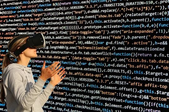Frau mit aufgesetzter Datenbrille steht mit leicht ausgestreckten Händen vor einer schwarten Wand, die mit Programmiercode vollgeschrieben ist.